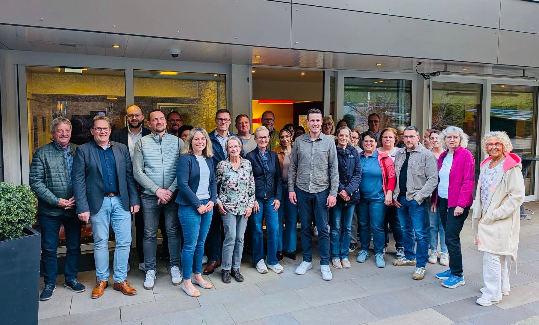 A group of about 23 adults stands together outside a building, smiling at the camera. They are casually dressed and appear to be participating in a group event or gathering.